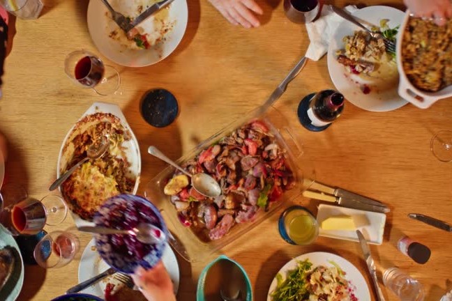 Overhead shot of a people gathered around a dining table loaded with food at a Friendsgiving potluck dinner eating and passing plates to one another.